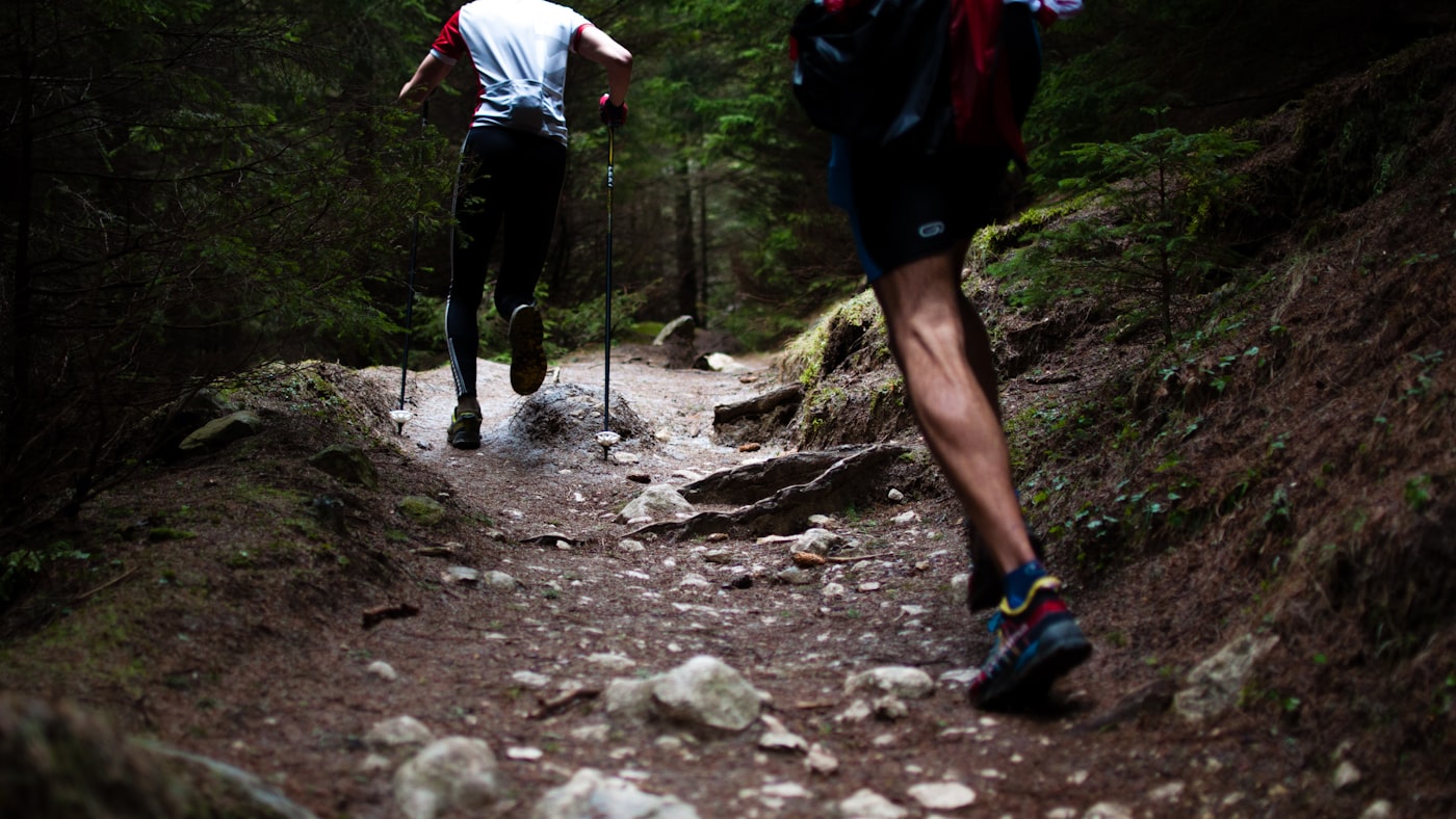 Two trail runners descending a rocky, root-covered path — the uneven terrain that rolls ankles and the reason balance training matters after an ankle sprain.
